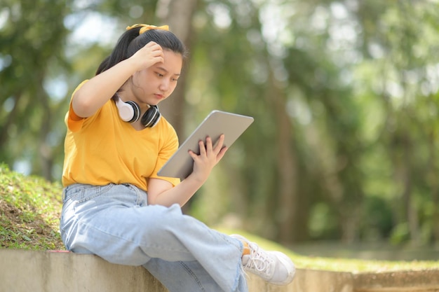 Adolescente de camisa amarela com fones de ouvido no pescoço segurando um tablet sentada no jardim