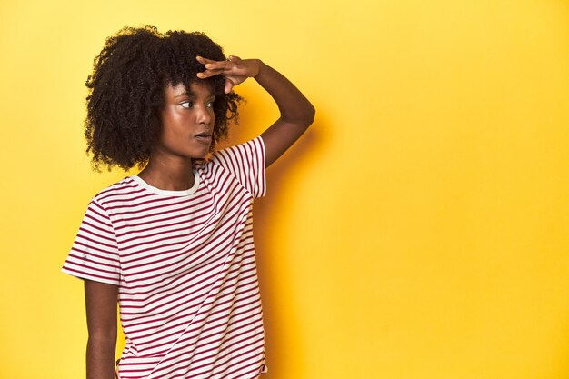 Adolescente de camiseta listrada vermelha com fundo amarelo de estúdio olhando para longe mantendo a mão na testa