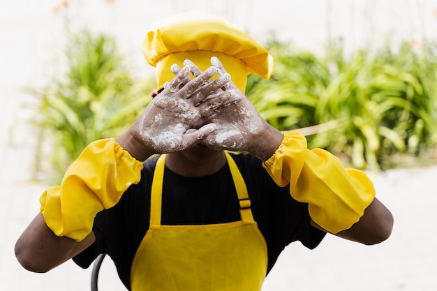 Adolescente de cozinheiro africano negro mostrando as mãos com farinha e fechando o rosto Criança africana com chapéu de chef e uniforme de avental amarelo
