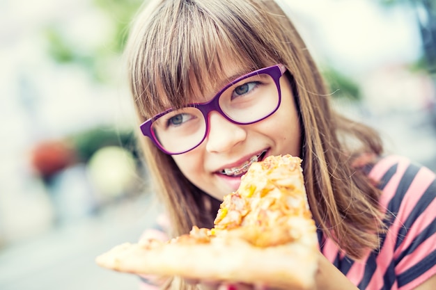 Adolescente de criança menina com pizza. Retrato de uma jovem pré-adolescente com um pedaço de pizza. Criança com aparelho e óculos. Foto tonificada.