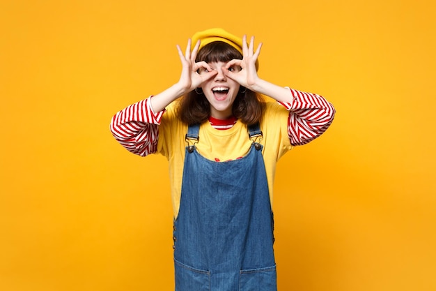 Adolescente de engraçadinha em vestido de denim de boina francesa segurando as mãos perto de olhos imitando óculos ou binóculos isolados em fundo amarelo. Conceito de estilo de vida de emoções sinceras de pessoas. Mock up espaço de cópia.