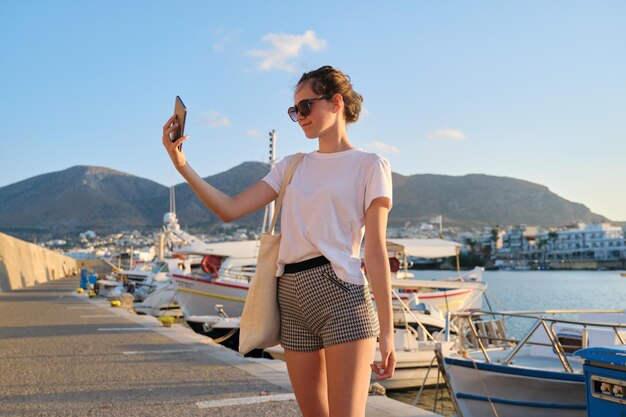 Adolescente de menina bonita elegante caminhando no cais, pôr do sol no mar, iates atracados na baía, fundo de paisagem de montanha. Garota de óculos escuros, shorts leva selfie no smartphone.