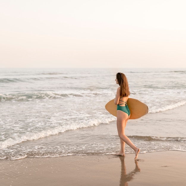 Foto adolescente delgada caminando cerca de la costa con tabla de surf en la playa