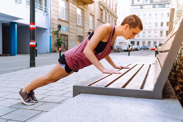 Una adolescente deportista hace flexiones con los brazos doblados en los codos desde un banco en las calles de la ciudad bajo el sol