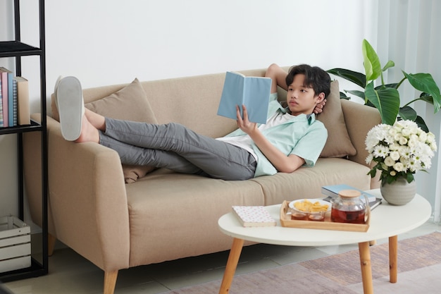 Adolescente descansando no sofá, bebendo chá e lendo um livro interessante com romances