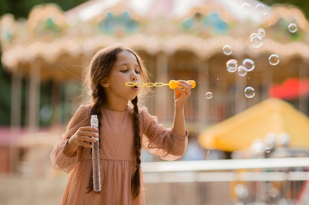Adolescente con dos coletas lanza burbujas en el parque de atracciones cerca del carrusel y se regocija