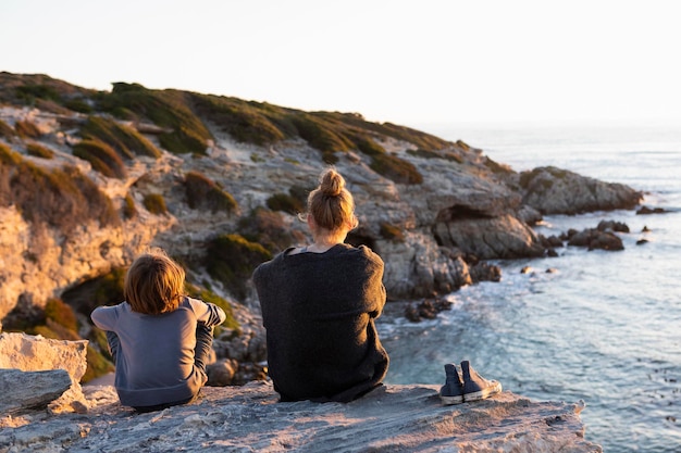 Adolescente e menino sentado nas rochas olhando para o mar ao pôr do sol