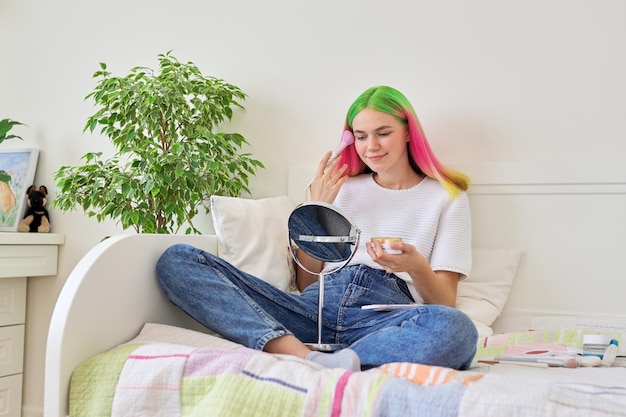 Adolescente elegante fazendo maquiagem em casa, garota com cabelo tingido na moda, sentado na cama com cosméticos e espelho. Beleza e maquiagem, moda e cosméticos na adolescência