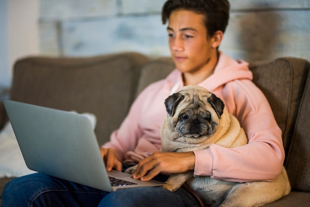 Adolescente em casa trabalhando com seu laptop no sofá com um pug ao lado dele - melhores amigos abraçados no sofá