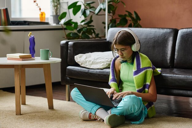 Adolescente em fones de ouvido sentado no chão na sala de estar e estudando online no laptop