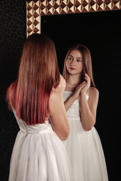 Adolescente em pé e posando olhando para o reflexo Retrato linda garota adolescente em um elegante vestido branco no espelho