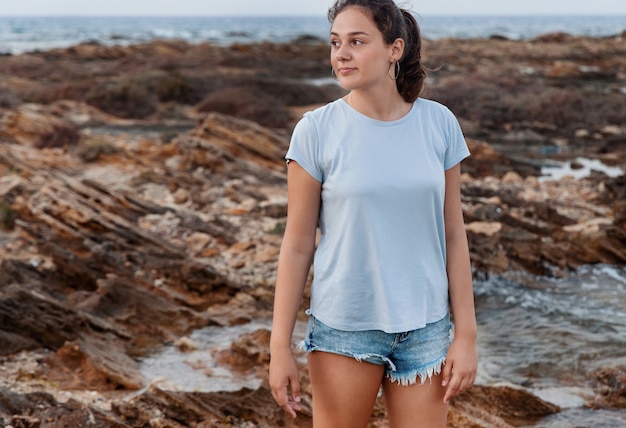 Adolescente em pé na falésia à beira-mar ao pôr do sol vestindo camiseta azul claro, shorts jeans e olhando de lado. tiro de comprimento de três quartos. maquete de camiseta