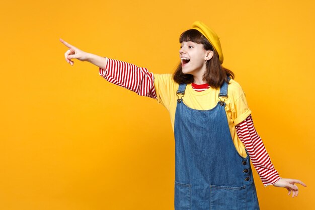 Adolescente emocionada con boina francesa, vestido de mezclilla apuntando con el dedo índice a un lado y manteniendo la boca abierta aislada en un fondo amarillo. Emociones sinceras de la gente, concepto de estilo de vida. Simulacros de espacio de copia.