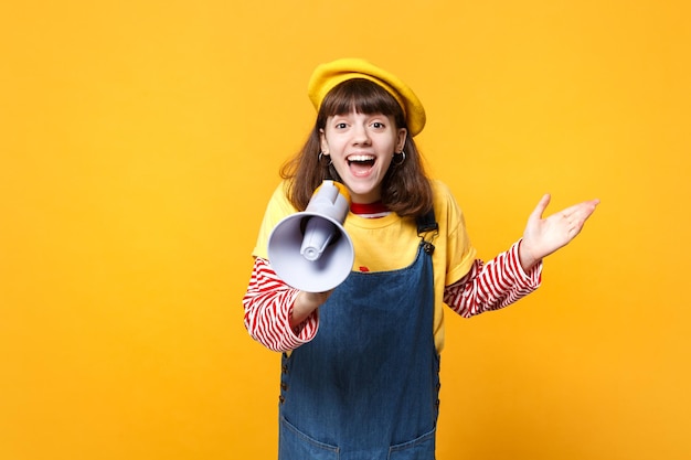 Adolescente emocionada con boina francesa, vestido de mezclilla gritando en megáfono, extendiendo las manos aisladas en el fondo de la pared amarilla en el estudio. Emociones sinceras de la gente, concepto de estilo de vida. Simulacros de espacio de copia.