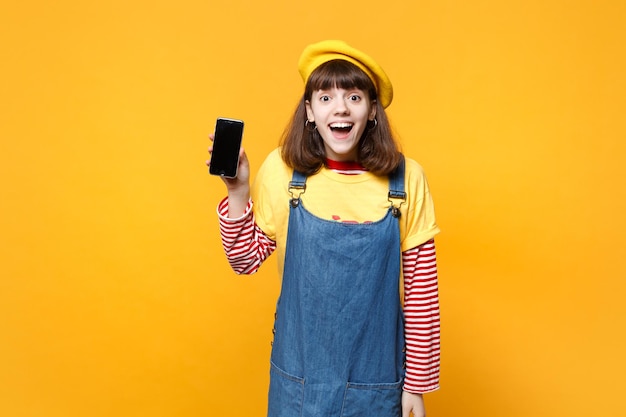 Adolescente emocionada con boina francesa, vestido de mezclilla sosteniendo un teléfono móvil con una pantalla vacía en blanco aislada en el fondo de la pared amarilla. Emociones sinceras de la gente, concepto de estilo de vida. Simulacros de espacio de copia.