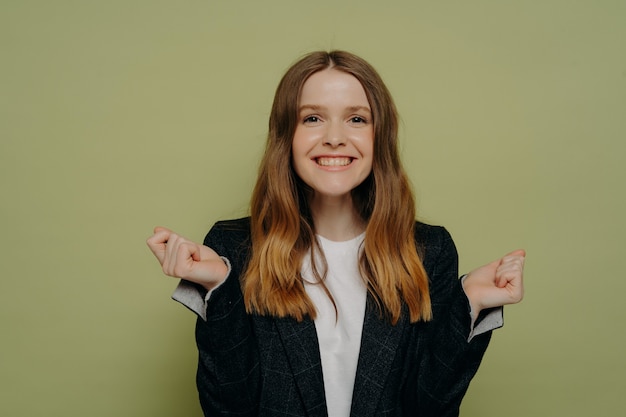 Adolescente emocionada con cabello ondulado en ropa casual celebrando la victoria y el éxito, siendo muy feliz, adolescente sonriente haciendo gesto de ganador mientras está de pie aislado sobre fondo verde