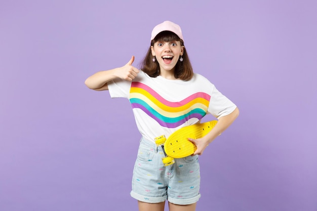 Una adolescente emocionada con ropa vívida con patineta amarilla, mostrando el pulgar hacia arriba, manteniendo la boca abierta aislada en un fondo violeta pastel. Emociones sinceras de la gente, concepto de estilo de vida. Simulacros de espacio de copia.