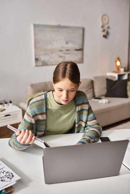 Adolescente enfocada con el aprendizaje electrónico de portátil con portátil cerca de bocetos y teléfono inteligente en el escritorio