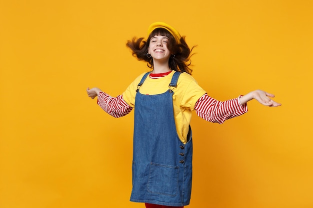 Adolescente engraçadinha na boina francesa, vestido jeans espalhando as mãos com cabelo solto isolado no fundo da parede amarela no estúdio. Emoções sinceras de pessoas, conceito de estilo de vida. Mock up espaço de cópia.