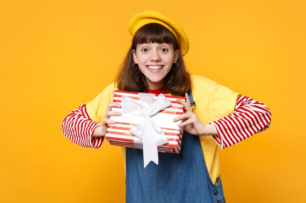 Adolescente engraçadinha na boina francesa, vestido jeans segurando a caixa de presente listrada vermelha com fita de presente isolada no fundo da parede amarela. Emoções sinceras de pessoas, conceito de férias de aniversário de estilo de vida.