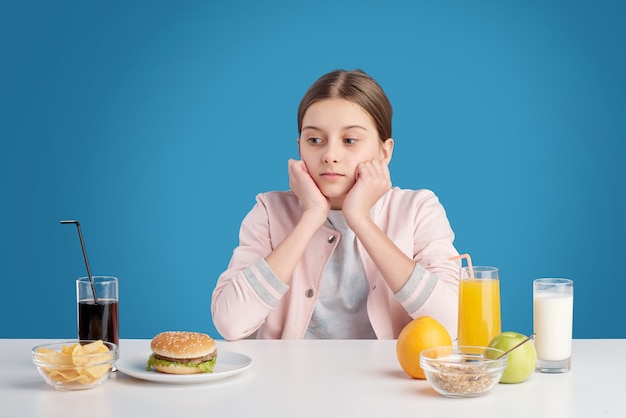 Adolescente escolhendo entre uma alimentação saudável orgânica fresca e junk food enquanto está sentada à mesa em frente à câmera, isolada