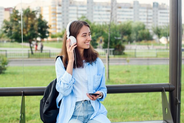 Adolescente escucha la música con auriculares blancos en una estación de transporte público mientras espera el tranvía.