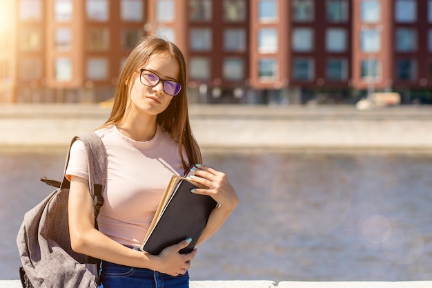 Adolescente estudante caminha pela cidade em óculos com livros e uma mochila em um dia de verão perto do rio