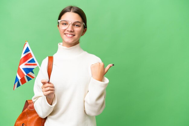 Adolescente estudiante chica caucásica sosteniendo una bandera del Reino Unido sobre un fondo aislado apuntando hacia un lado para presentar un producto