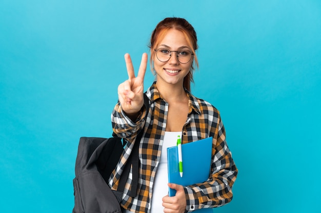 Adolescente estudiante chica rusa aislada en la pared azul sonriendo y mostrando el signo de la victoria