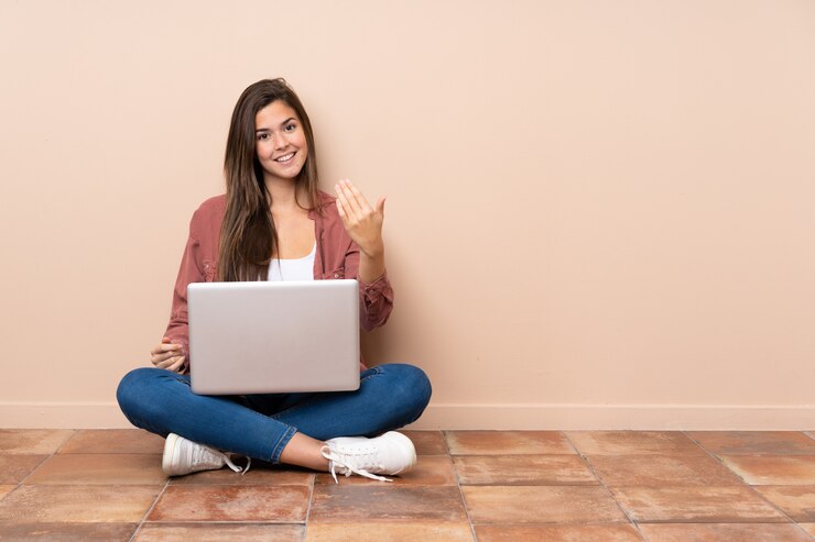 Foto adolescente estudiante mujer sentada en el suelo con un portátil invitando a venir con la mano. feliz de que hayas venido