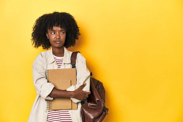 Foto adolescente estudiosa con mochila y cuadernos telón de fondo amarillo confundida se siente dudosa e insegura