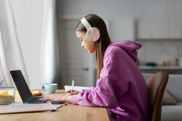 Una adolescente europea tiene una lección en línea usando una computadora portátil con auriculares tomando notas durante la línea