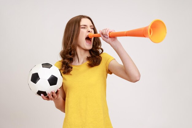 Una adolescente extremadamente positiva y satisfecha con una camiseta amarilla gritando con megáfono sosteniendo una pelota de fútbol, celebrando la victoria de su equipo de fútbol favorito. Disparo de estudio interior aislado sobre fondo gris.