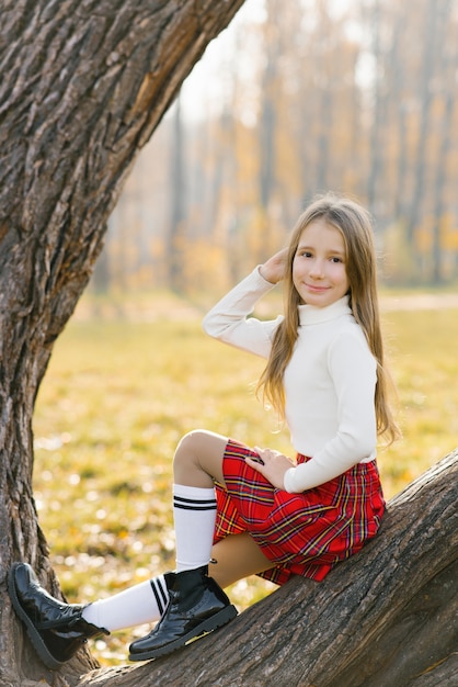 Una adolescente con una falda roja y un suéter blanco se sienta en un árbol en un parque de otoño y sonríe