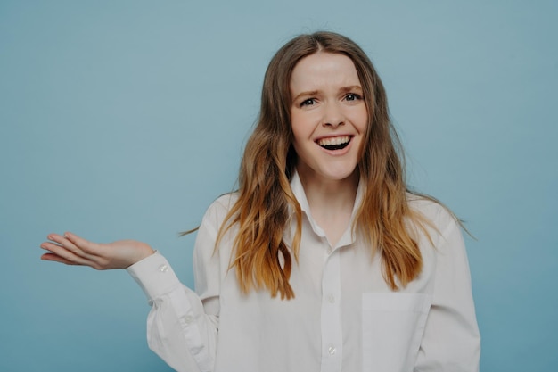 Una adolescente feliz con camisa blanca que presenta algo en la mano con la palma levantada demostrando un producto publicitario aislado en el fondo azul
