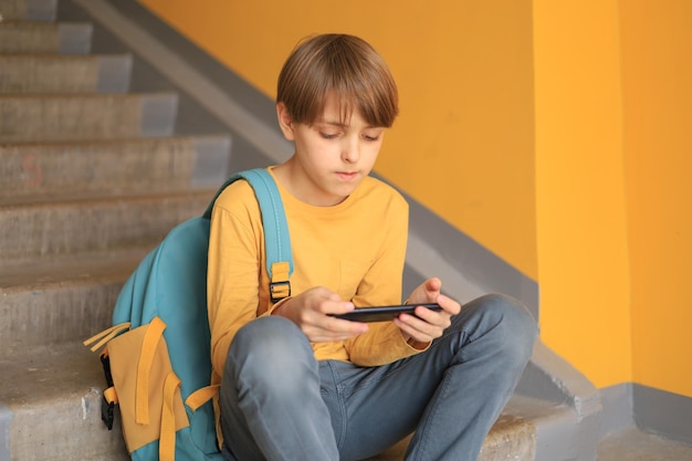 Un adolescente feliz con una camiseta amarilla se sienta con un teléfono cerca de la pared amarilla. Usando tecnología en línea