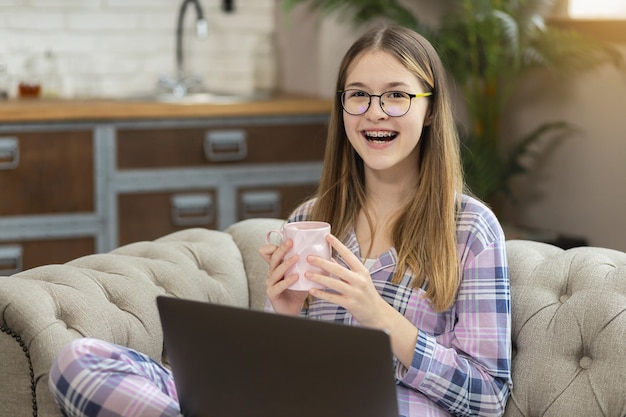 Adolescente feliz com um sorriso de pijama e um laptop segurando uma xícara de chá e olhando diretamente para a câmera enquanto está sentado no sofá em casa