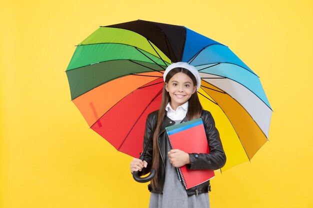 Una adolescente feliz con gafas y boina bajo un paraguas colorido para protegerse de la lluvia en la temporada de otoño tiene educación portátil