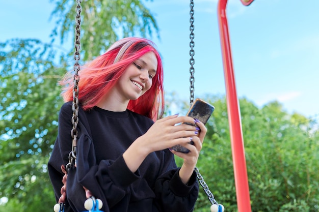 Adolescente feliz de moda con cabello teñido con columpio de equitación de teléfono inteligente