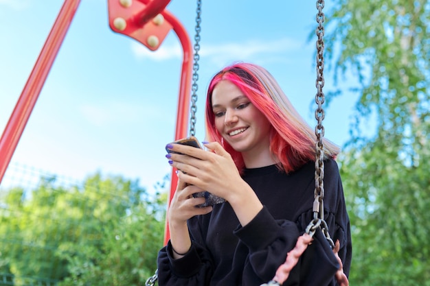 Adolescente feliz de moda con cabello teñido con teléfono inteligente montando columpio, escribiendo mensajes de texto, comunicándose en redes sociales, blogueando por teléfono. Estilo de vida adolescente, tecnología, concepto de juventud.