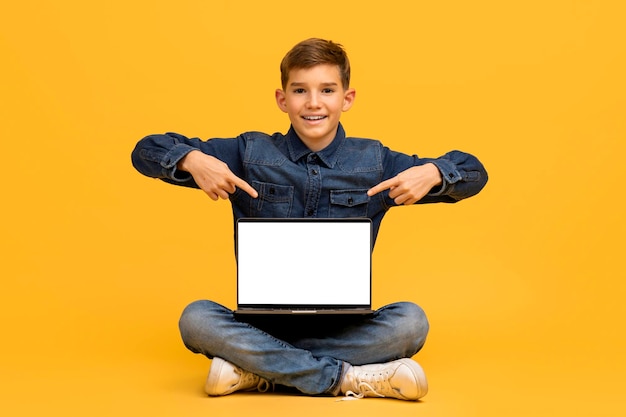 Adolescente feliz sentado en el suelo señalando la pantalla en blanco de un portátil