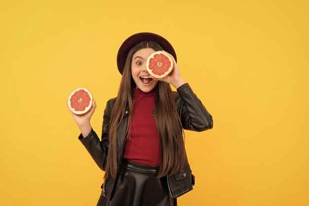 Una adolescente feliz con sombrero y chaqueta de cuero sostiene alimentos saludables de pomelo cortado