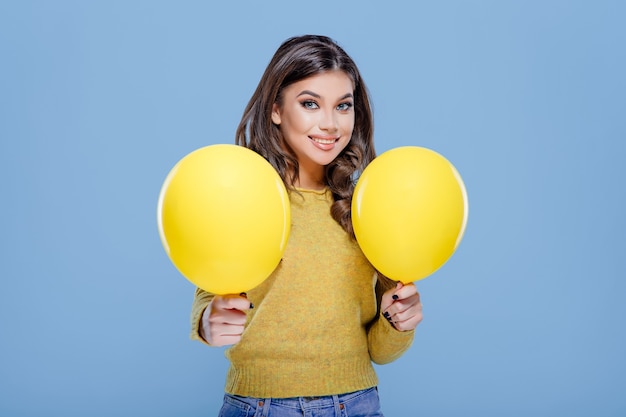 Adolescente feliz sosteniendo globos amarillos en la mano y sonriendo para la cámara mientras se divierte contra b ...