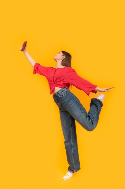 Adolescente feliz con teléfono sobre fondo amarillo brillante Retrato completo de una joven en jeans y camiseta roja toma fotos de sí misma