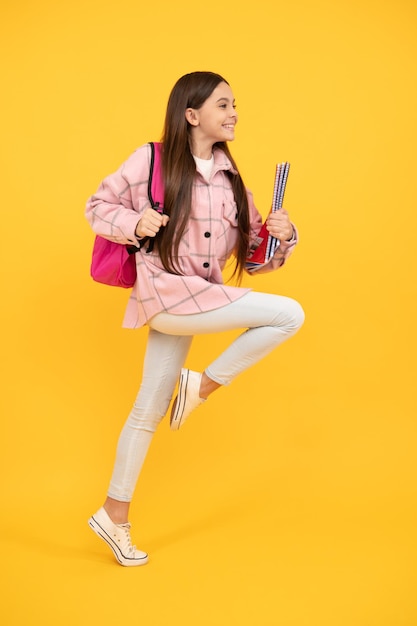 una adolescente feliz usa una camisa rosa a cuadros corriendo con una mochila escolar y cuadernos, corredora.