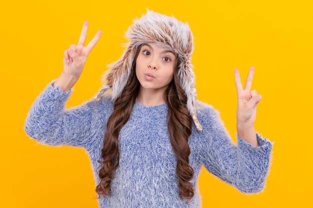 Una adolescente feliz usa un sombrero con orejeras que muestra un gesto de paz sobre fondo amarillo, invierno