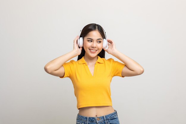 Adolescente feminino asiático alegre ouve a música com fone de ouvido branco isolado. Mulher jovem e bonita na mão da camisa amarela toca um fone de ouvido sem fio se divertindo com a música no quarto.