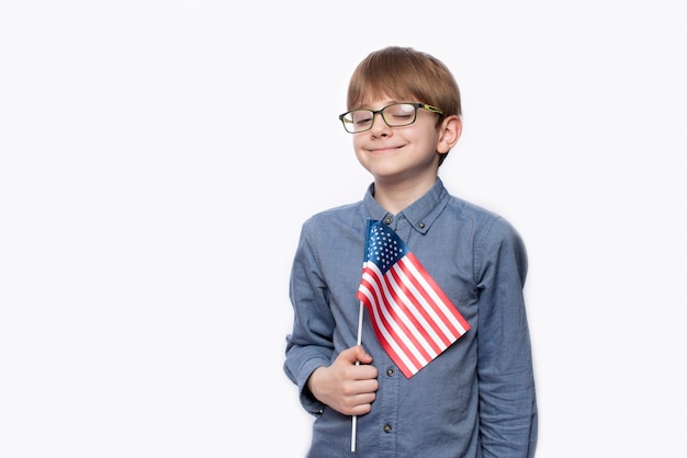 Adolescente fofo sério de óculos com a bandeira americana na parede branca isolada