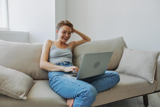 Adolescente freelancer com laptop sentada no sofá em casa sorrindo com roupas de casa e óculos com corte de cabelo curto estilo de vida sem filtros espaço de cópia livre foto de alta qualidade
