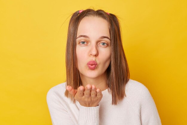 Adolescente funky romântica com rabos de cavalo em suéter posando isolado sobre fundo amarelo enviando ar beijando flertando com o namorado mantém os lábios fazendo beicinho olhando para a câmera